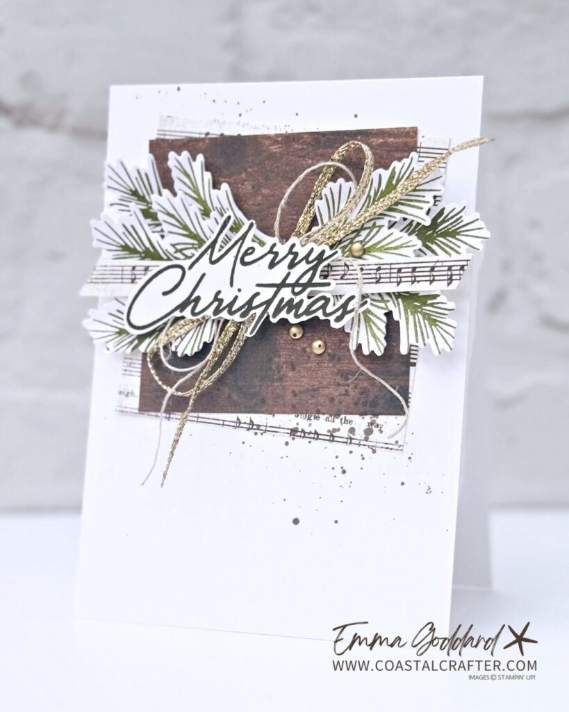 Handmade Christmas card on white cardstock. In the centre, a dark wood grain panel is layered over strips of vintage sheet music, topped with a cluster of die cut pine branches stamped in olive green. A "Merry Christmas" sentiment in dark script sits across the middle, with loops of gold metallic thread and natural twine, three small gold embellishments, and soft grey ink splatters around the design.