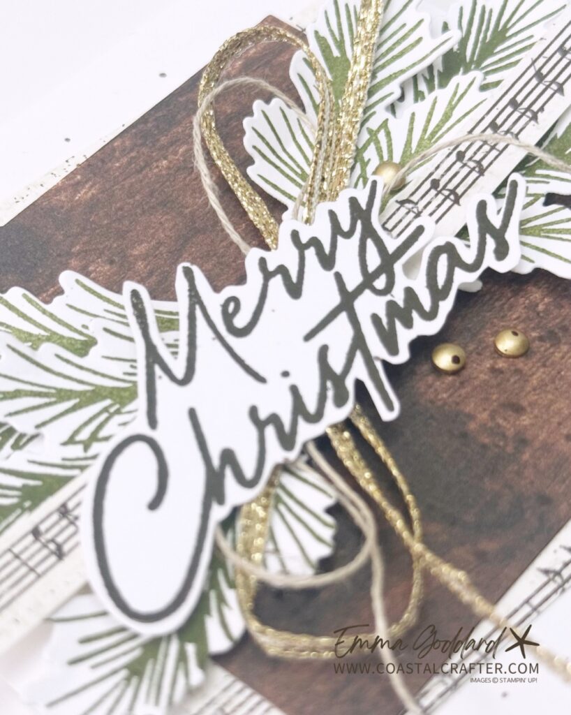 Handmade Christmas card on white cardstock. In the centre, a dark wood grain panel is layered over strips of vintage sheet music, topped with a cluster of die cut pine branches stamped in olive green. A "Merry Christmas" sentiment in dark script sits across the middle, with loops of gold metallic thread and natural twine, three small gold embellishments, and soft grey ink splatters around the design.