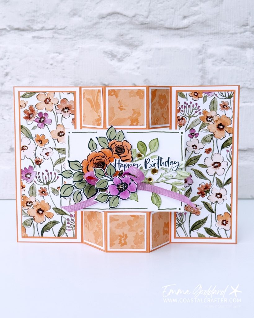 A handmade floral birthday card with a unique gatefold design. The card features panels decorated with watercolour orange, purple, and white flowers on a white background. The centre panel displays a cluster of coloured flowers in orange and purple, accented with green leaves, a purple ribbon, and a "Happy Birthday" sentiment in elegant script. The card has a dimensional, layered look and is framed in timid tiger cardstock. The creator's watermark, "Emma Goddard | www.coastalcrafter.com," is visible in the bottom right corner.
