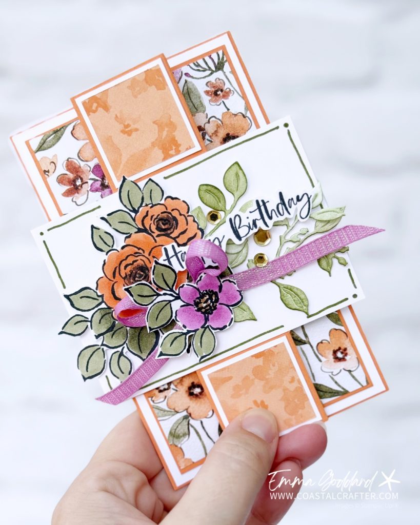 A handmade floral birthday card with a unique gatefold design. The card features panels decorated with watercolour orange, purple, and white flowers on a white background. The centre panel displays a cluster of coloured flowers in orange and purple, accented with green leaves, a purple ribbon, and a "Happy Birthday" sentiment in elegant script. The card has a dimensional, layered look and is framed in timid tiger cardstock. The creator's watermark, "Emma Goddard | www.coastalcrafter.com," is visible in the bottom right corner.