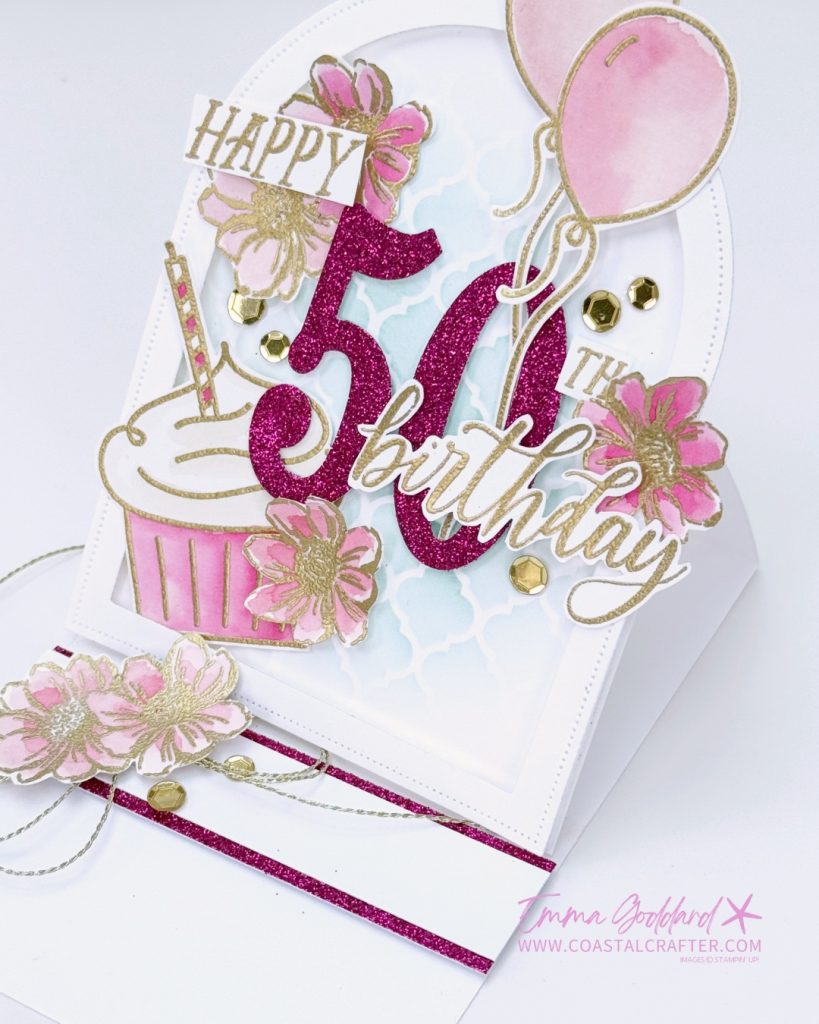 Handmade 50th birthday card in pink, white, and gold tones. The card features a glittery pink number "50" with "Happy" and "Birthday" written in gold script. Decorative elements include pink watercolour flowers, gold sequins, pink balloons, a cupcake, and a soft blue stencil background. The card is arranged in an easel style with layered embellishments and a glittery pink strip at the base. Designed by Emma Goddard, as indicated by the watermark.