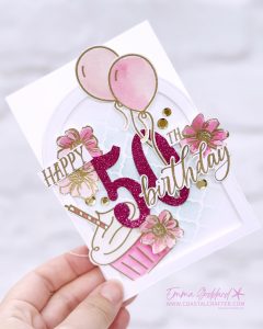 Handmade 50th birthday card in pink, white, and gold tones. The card features a glittery pink number "50" with "Happy" and "Birthday" written in gold script. Decorative elements include pink watercolour flowers, gold sequins, pink balloons, a cupcake, and a soft blue stencil background. The card is arranged in an easel style with layered embellishments and a glittery pink strip at the base. Designed by Emma Goddard, as indicated by the watermark.