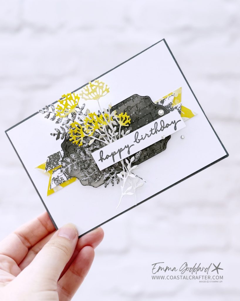 A handmade birthday card held in a person's hand, featuring layered die-cut floral elements in yellow, white, and gray tones. The card has a white background with a dark gray border and a central decorative label shape in textured dark gray with script text. Yellow and patterned gray banner strips peek out from behind the label. The sentiment “happy birthday” is stamped in a modern handwritten style on a white strip. Embellishments include small adhesive pearls. The creator’s name, Emma Goddard, and website www.coastalcrafter.com are printed in the bottom right corner.