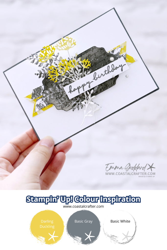 A handmade birthday card featuring die-cut flowers in yellow (Darling Duckling), white (Basic White), and gray (Basic Gray) layered over a textured dark gray label with a "happy birthday" sentiment in script. The card uses a minimal and elegant design with white background and accent banners. Below the card, there’s a color palette labeled “Stampin’ Up! Colour Inspiration” showing three swatches: Darling Duckling (yellow), Basic Gray (gray), and Basic White (white). The creator’s name, Emma Goddard, and website www.coastalcrafter.com are included.
