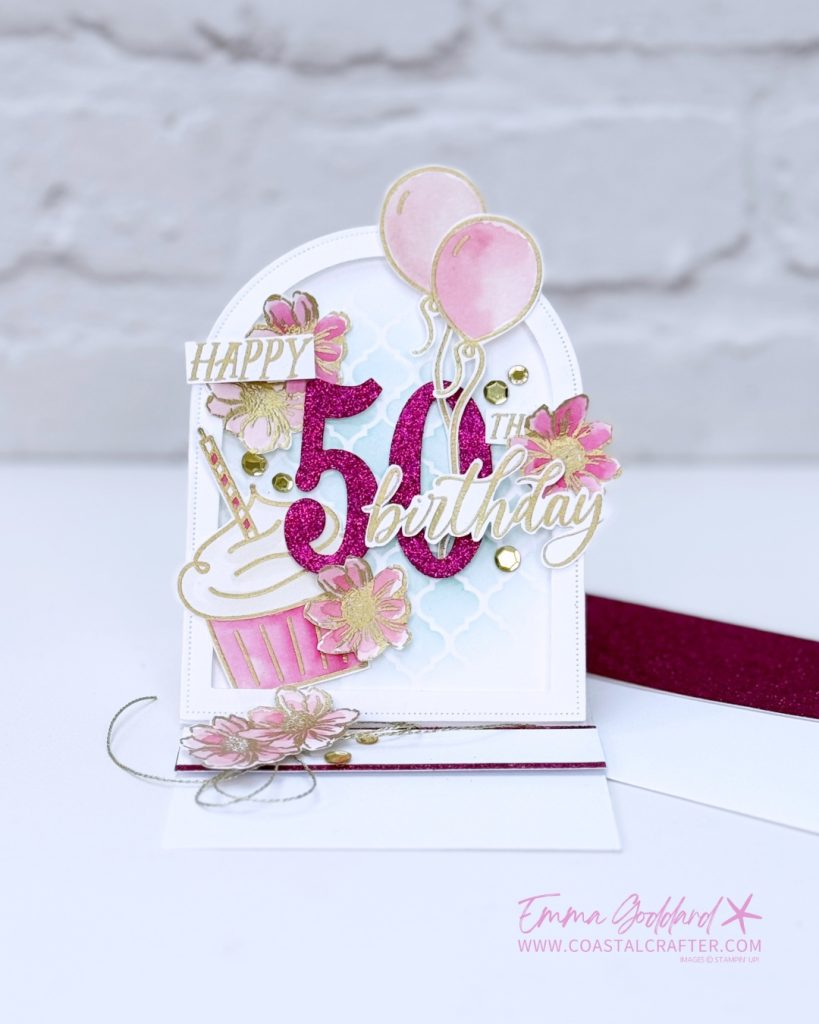 Handmade 50th birthday card in pink, white, and gold tones. The card features a glittery pink number "50" with "Happy" and "Birthday" written in gold script. Decorative elements include pink watercolour flowers, gold sequins, pink balloons, a cupcake, and a soft blue stencil background. The card is arranged in an easel style with layered embellishments and a glittery pink strip at the base. Designed by Emma Goddard, as indicated by the watermark.