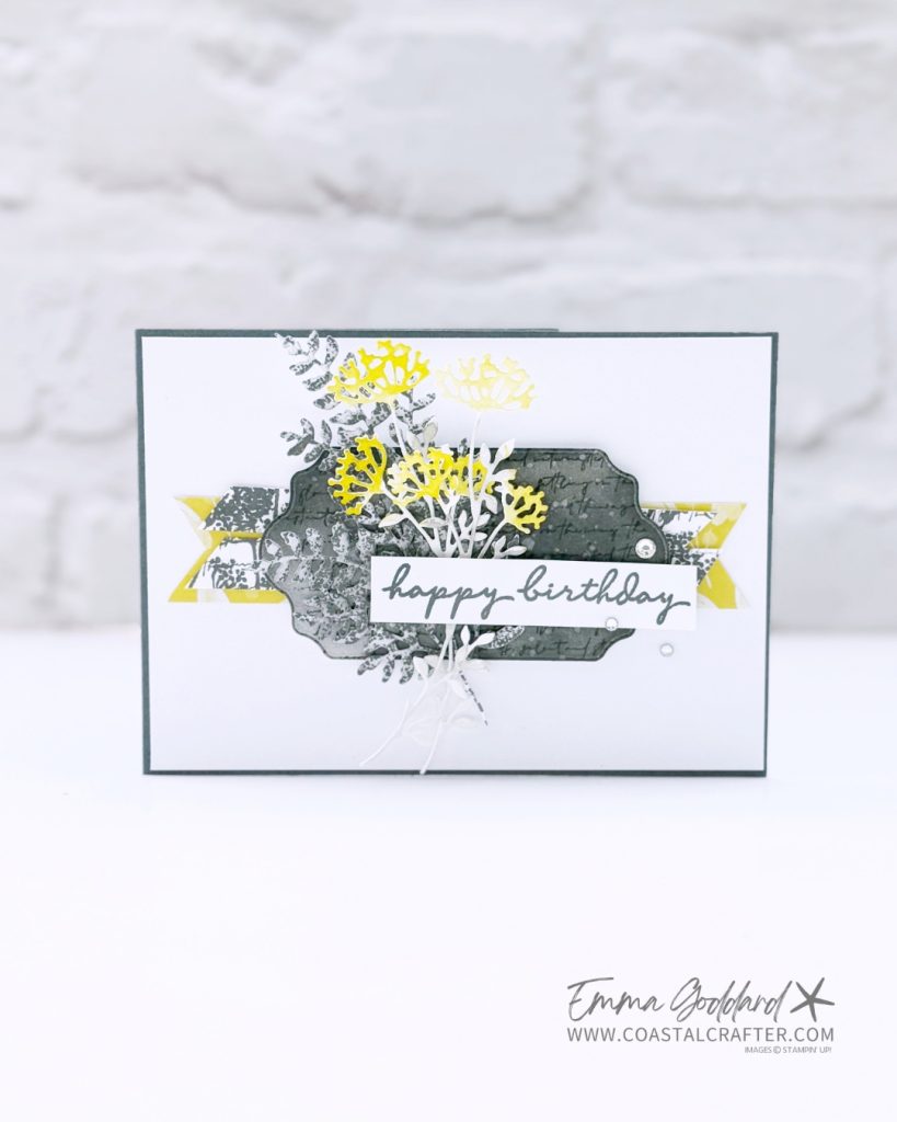 A handmade birthday card held in a person's hand, featuring layered die-cut floral elements in yellow, white, and gray tones. The card has a white background with a dark gray border and a central decorative label shape in textured dark gray with script text. Yellow and patterned gray banner strips peek out from behind the label. The sentiment “happy birthday” is stamped in a modern handwritten style on a white strip. Embellishments include small adhesive pearls. The creator’s name, Emma Goddard, and website www.coastalcrafter.com are printed in the bottom right corner.