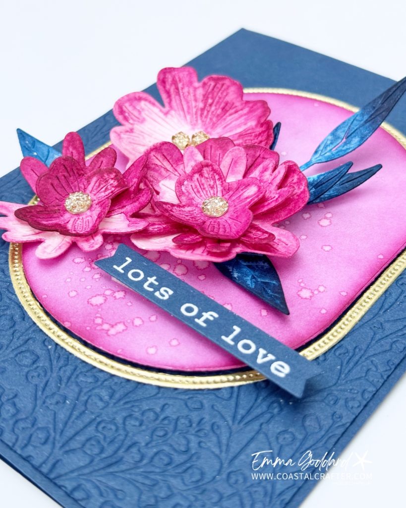 Handmade greeting card featuring vibrant, three-dimensional pink and magenta flowers with glittery gold centers, set against a pink oval background with gold trim. The card has a textured navy blue base with embossed floral patterns and a sentiment strip that reads "lots of love" in white text. Blue paper leaves accent the floral arrangement. The card is signed by Emma Goddard from coastalcrafter.com.