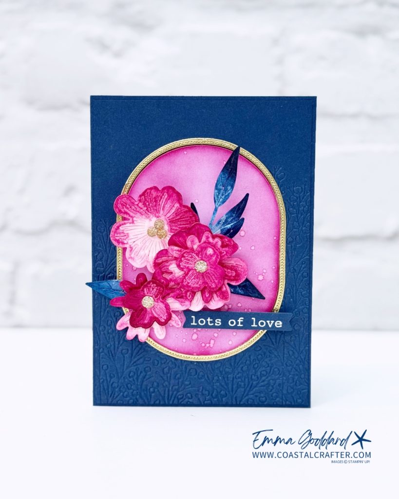 Handmade greeting card featuring vibrant, three-dimensional pink and magenta flowers with glittery gold centers, set against a pink oval background with gold trim. The card has a textured navy blue base with embossed floral patterns and a sentiment strip that reads "lots of love" in white text. Blue paper leaves accent the floral arrangement. The card is signed by Emma Goddard from coastalcrafter.com.